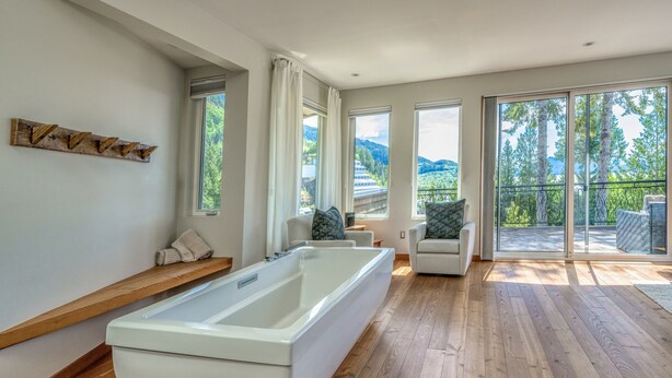 Een moderne badkamer met een groot bad, houten vloeren en grote ramen met uitzicht op bomen en bergen.