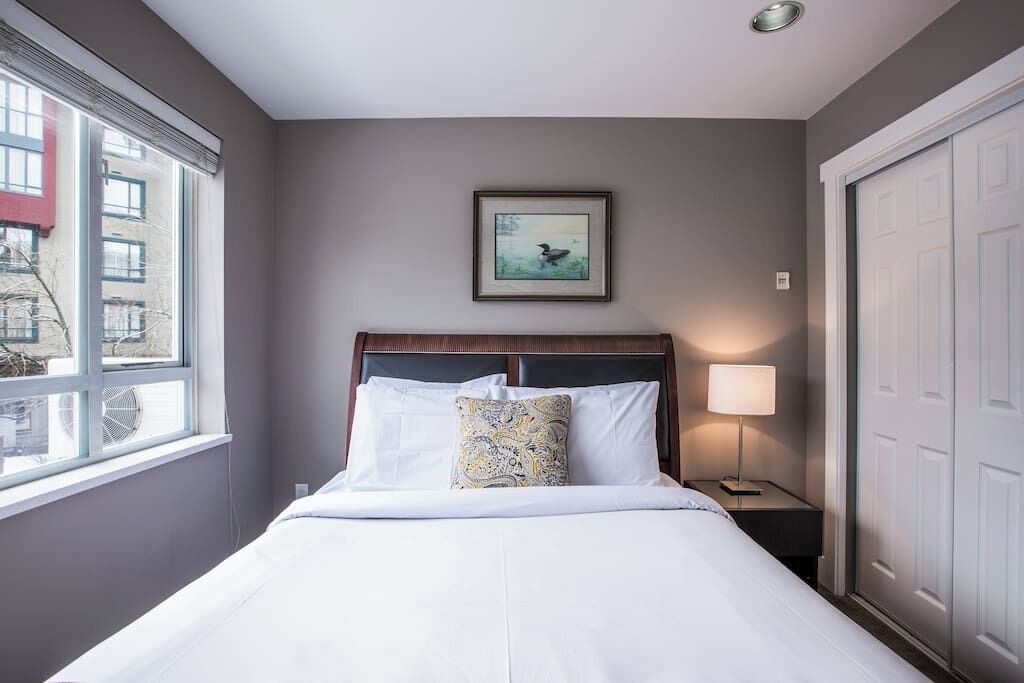 A hotel room with a bed, a bedside table with a lamp, a framed picture on the wall, and a window.