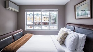 Habitación de hotel con un ventanal grande, cama con ropa blanca, una manta marrón, un cuadro en la pared y un equipo de aire acondicionado integrado.