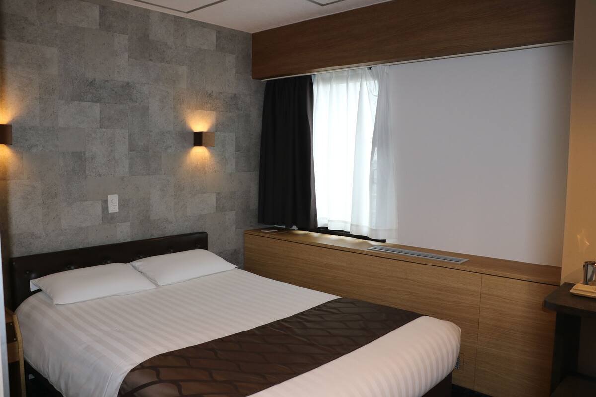 A modern hotel room with a large bed, a wooden headboard, a stone wall, and a window with curtains.