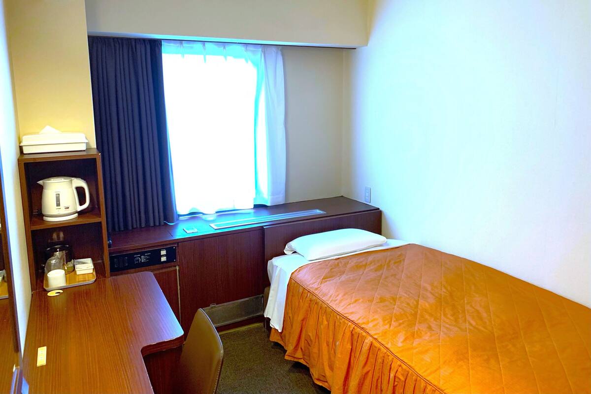 A hotel room with a bed, a desk with a kettle, a shelf with a kettle, a window with curtains, and a radiator.