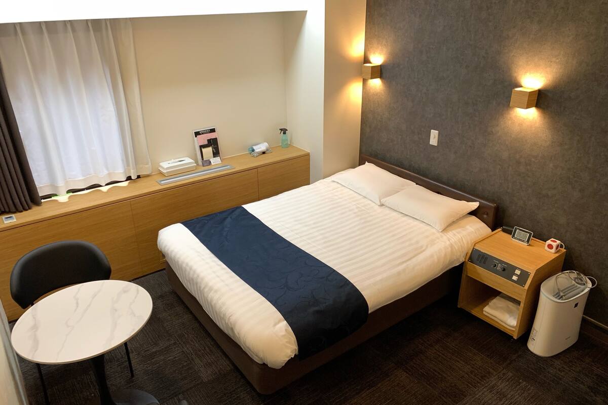A hotel room with a bed, bedside table, chair, desk, and a small table.