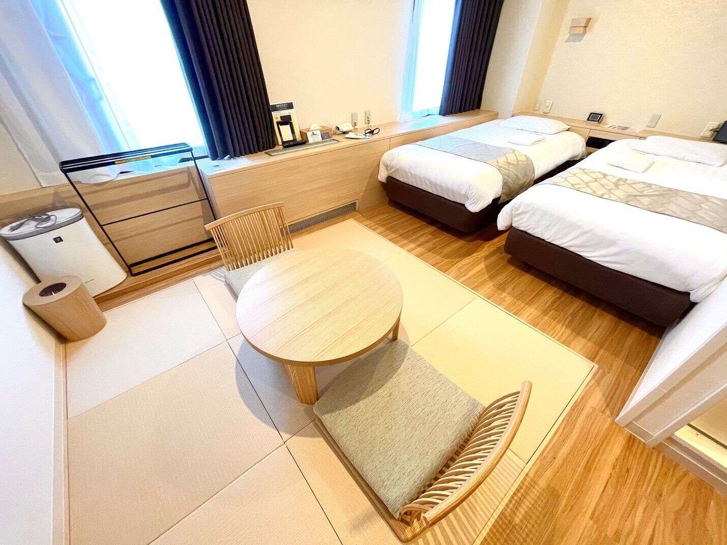 A hotel room with two beds, a small table, a chair, a desk with a computer, and a window with curtains.