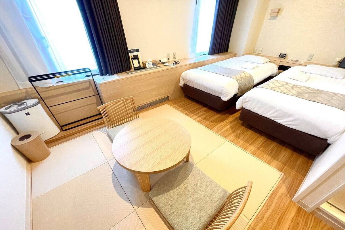 A hotel room with two beds, a small table, a chair, a desk with a computer, and a window with curtains.