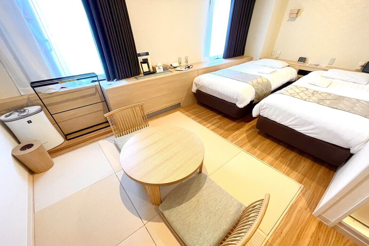 Habitación de hotel con dos camas, una mesita, una silla, un escritorio con computadora y una ventana con cortinas.