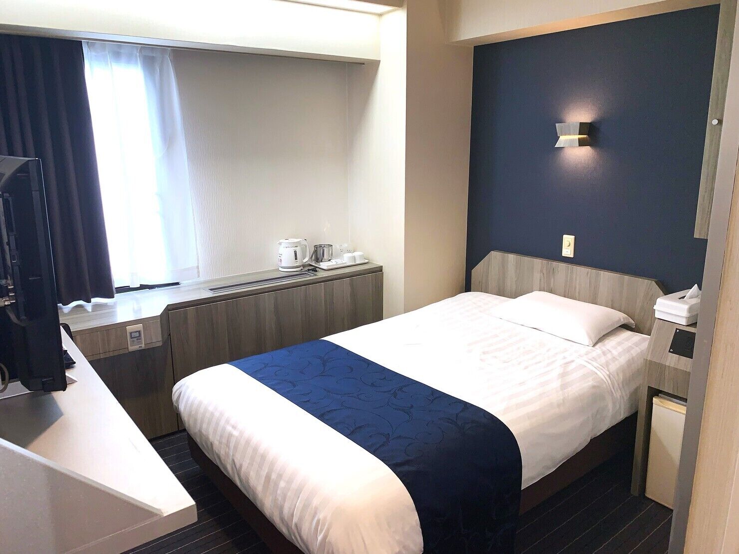 A hotel room with a bed, bedside table, a desk with a chair, a window with curtains, and a wall-mounted light fixture.