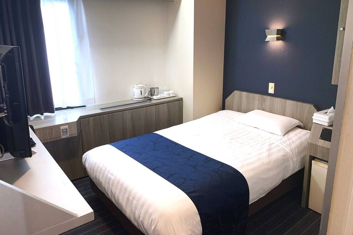 A hotel room with a bed, bedside table, a desk with a chair, a window with curtains, and a wall-mounted light fixture.