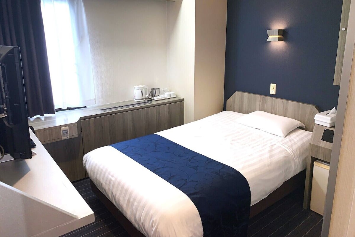 A hotel room with a bed, bedside table, a desk with a chair, a window with curtains, and a wall-mounted light fixture.