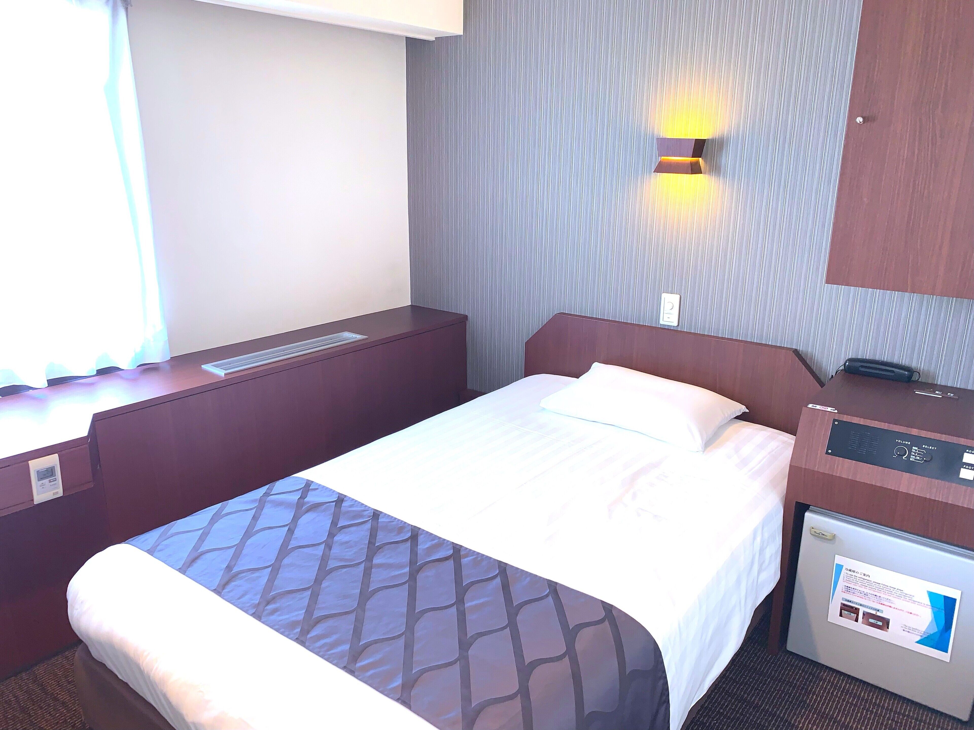 A hotel room with a bed, a desk, a wall-mounted lamp, and a control panel.
