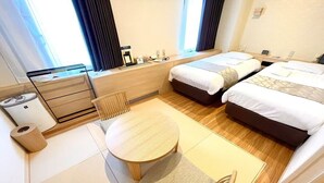 A hotel room with two beds, a small table, a waste bin, and a window with curtains.