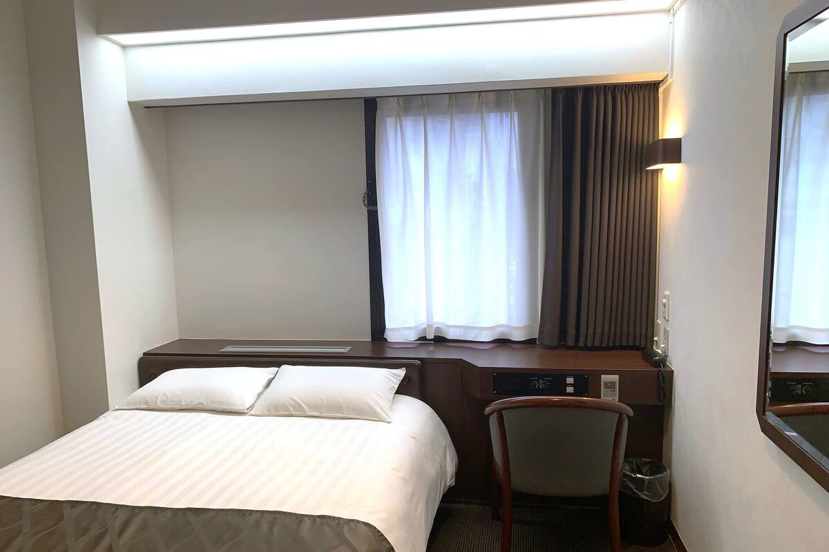 A hotel room with a bed, a desk, a chair, a window with curtains, and a mirror.