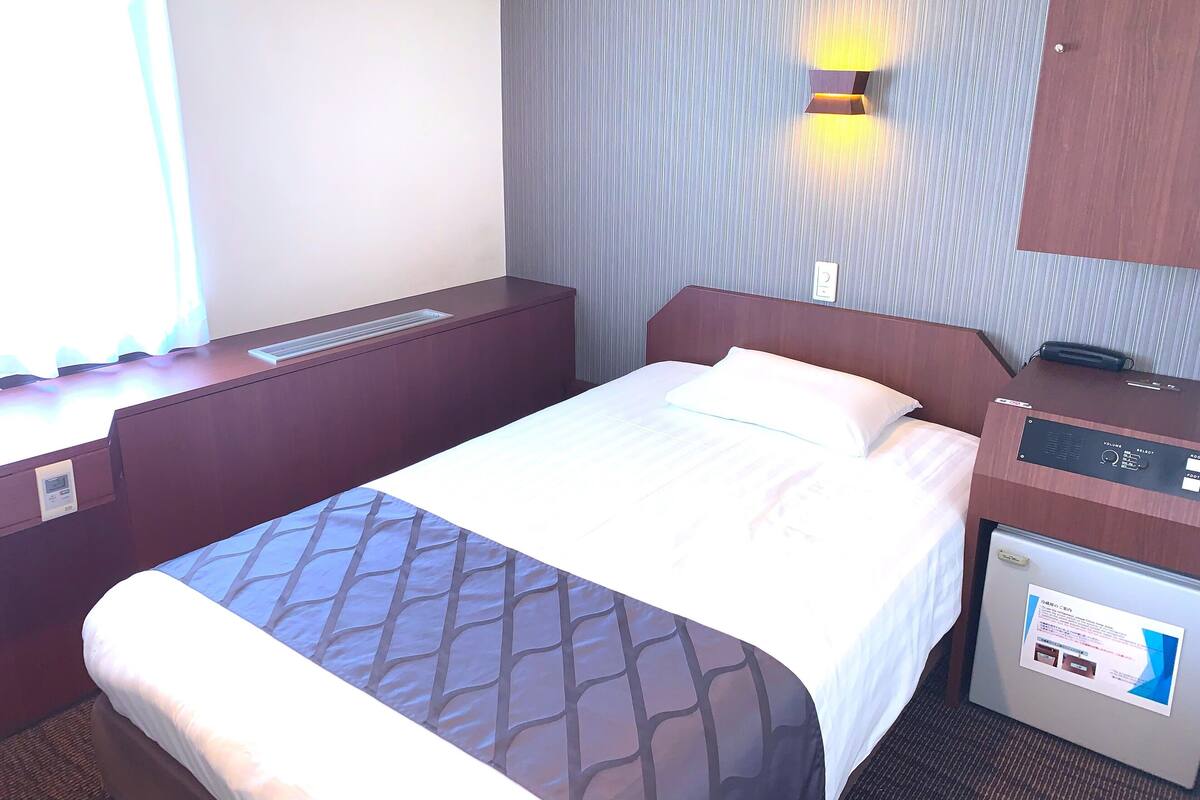 A hotel room with a bed, a desk, a chair, a control panel, and a wall-mounted light.