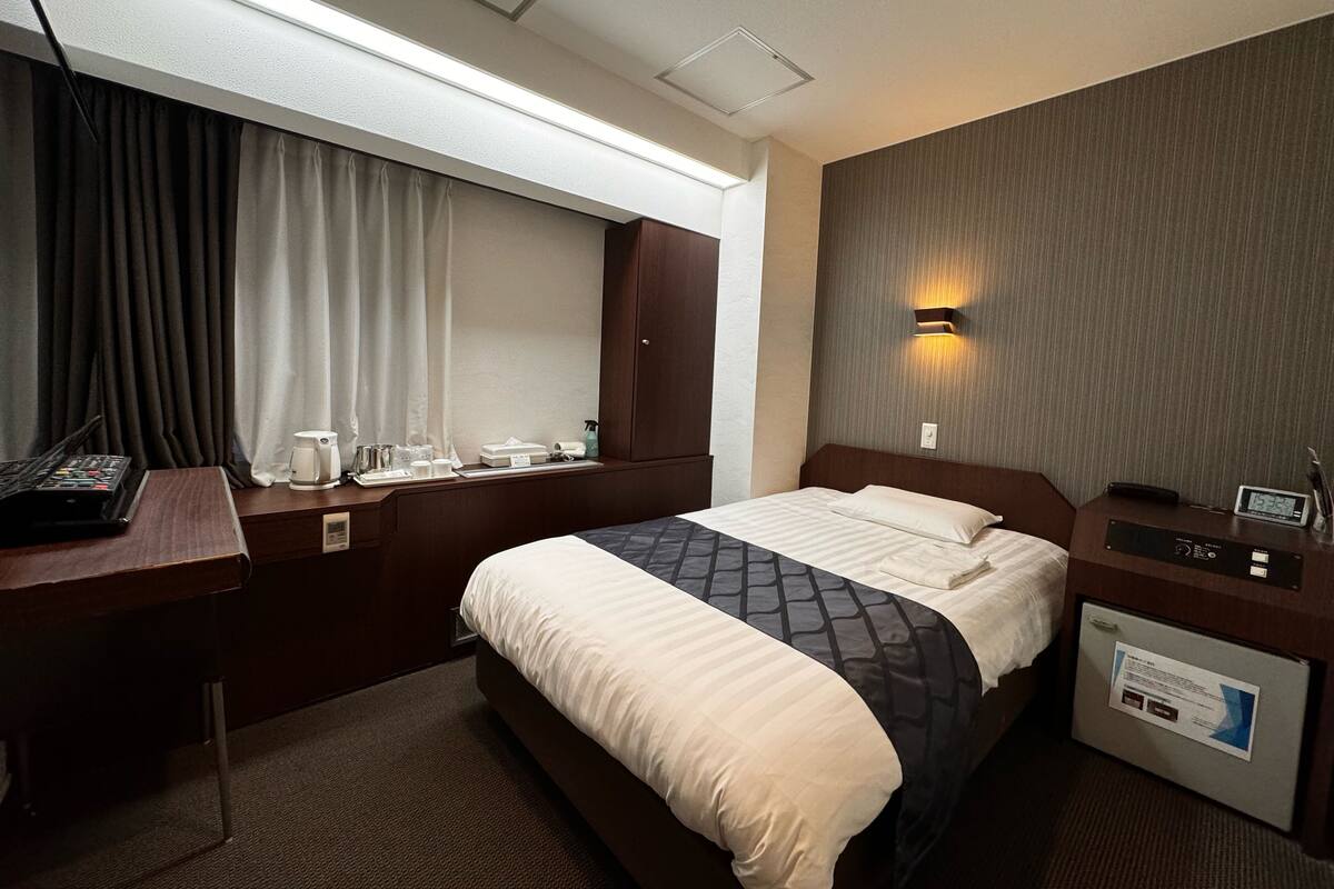 A hotel room with a bed, bedside table, desk, and a window with curtains.