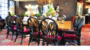A restaurant with a large dining table surrounded by chairs with red cushions. There is a chandelier hanging from the ceiling and a clock on the wall.