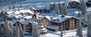 Ein schneebedecktes Resort mit mehreren Gebäuden, darunter ein markantes Logis mit einem Uhrturm und ein großes, rotes Gebäude mit Balkonen.