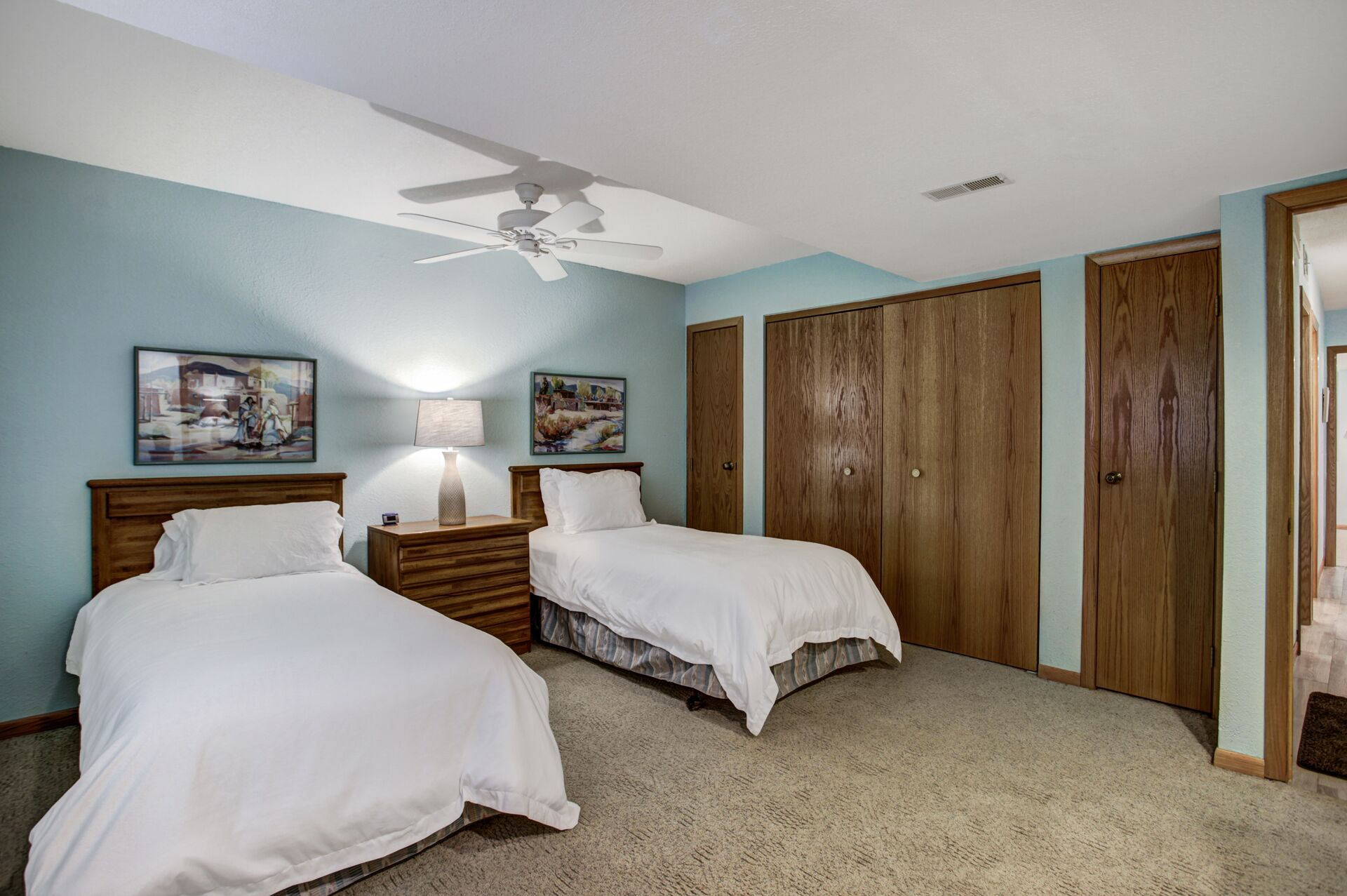 A bedroom with two beds, a ceiling fan, wooden wardrobes, and artwork on the walls.