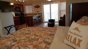 Ein Schlafzimmer mit einem Bett, einem Kopfkissen mit dem Aufdruck 'JimmyBahamand Relax', einer Küchenzeile mit Kühlschrank, Mikrowelle und Herd, einer Bar mit Barhocker und einer Glastür zum Außenbereich.