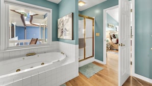 A modern bathroom with a large bathtub, a walk-in shower, and a view of a bedroom through an open door.