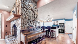 A spacious living area with a stone fireplace, a wooden piano, and a kitchen in the background.