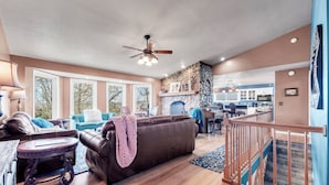 A spacious living room with a brown leather sofa, a round coffee table, and a ceiling fan. A kitchen is visible in the background with blue cabinets and a stone backsplash.
