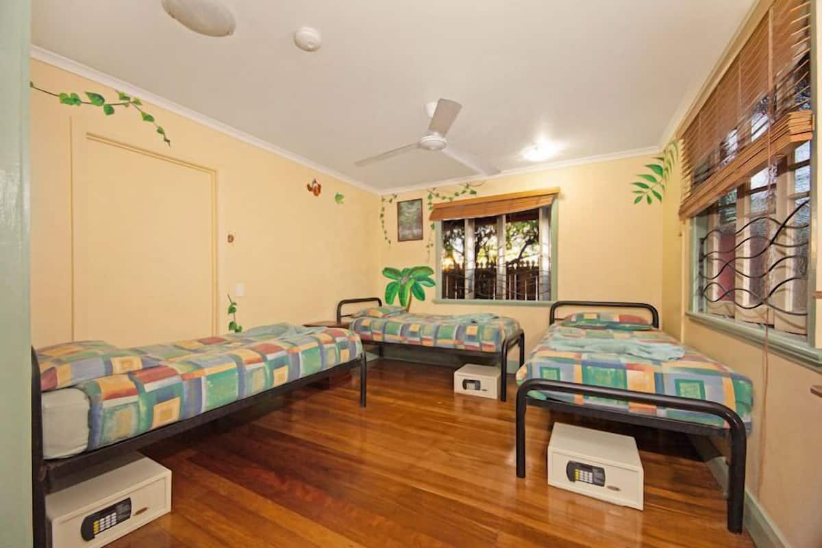 A room with three single beds, a ceiling fan, a window with blinds, and a door.