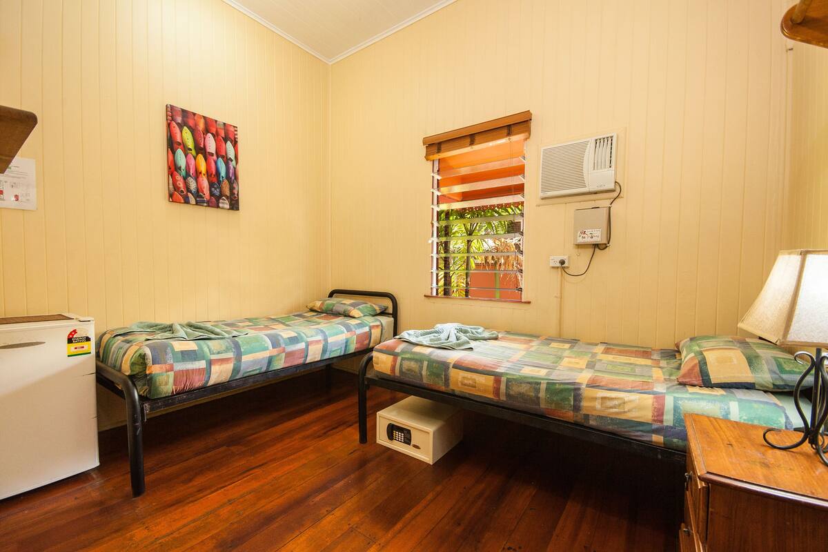 A room with two single beds, a small white refrigerator, a wooden cabinet, a colorful painting, and a window with blinds.