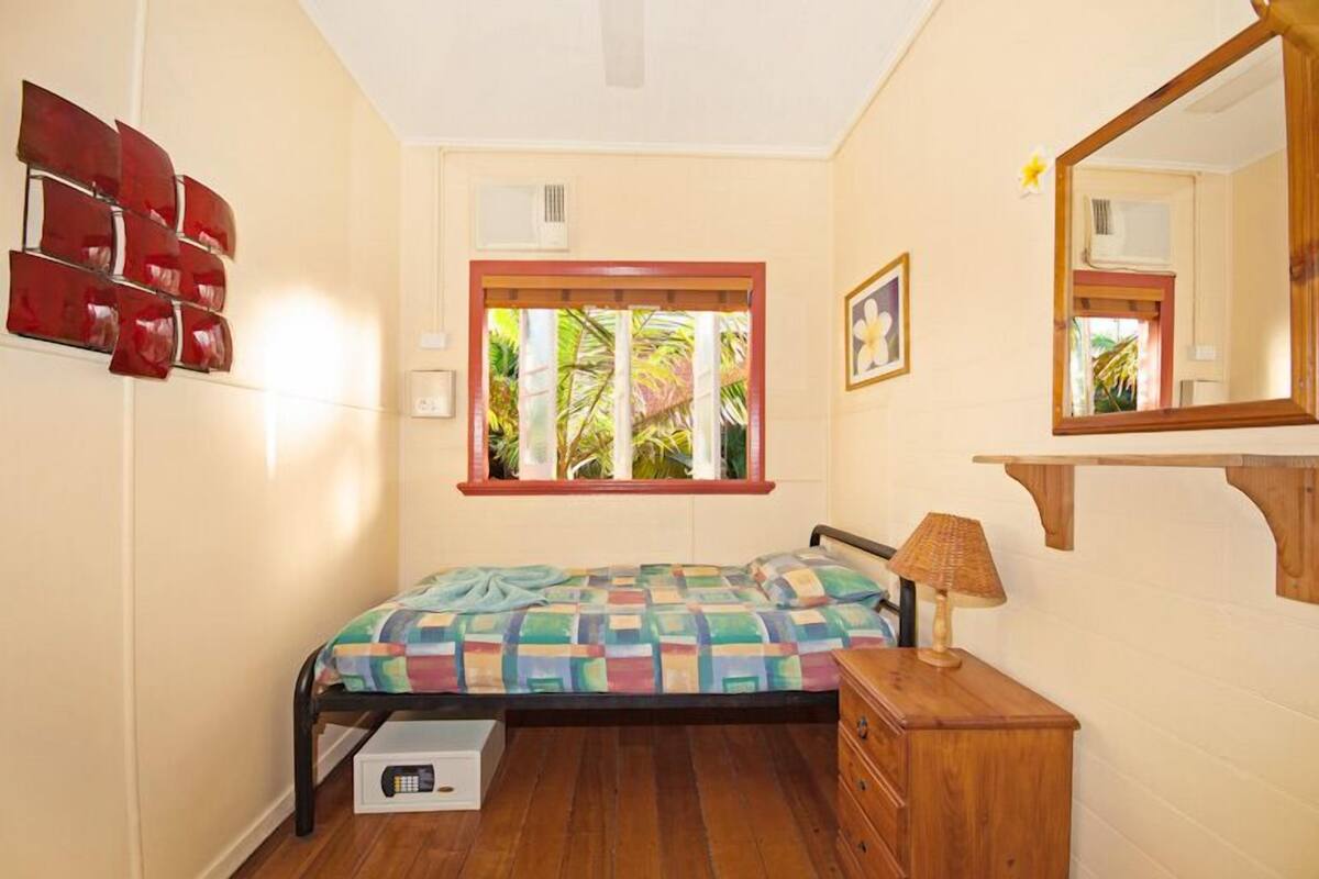 A bedroom with a bed, a wooden dresser, a window with a view of greenery, a wall-mounted mirror, and a red abstract art piece.
