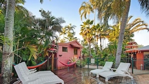 Une terrasse en bois avec des chaises longues blanches, une maison rose et des palmiers tout autour.