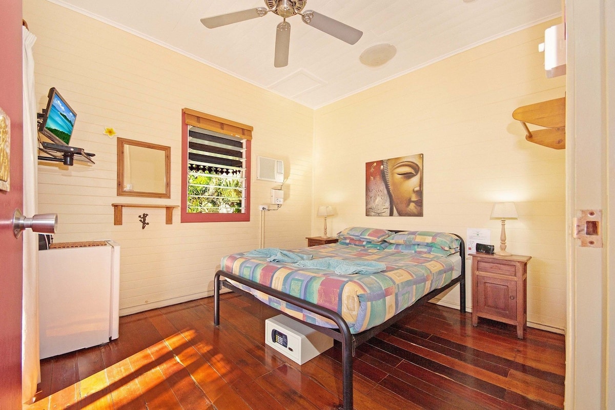 A bedroom with a bed, a ceiling fan, a window with blinds, a wall-mounted TV, a bedside table with a lamp, and a small cabinet.