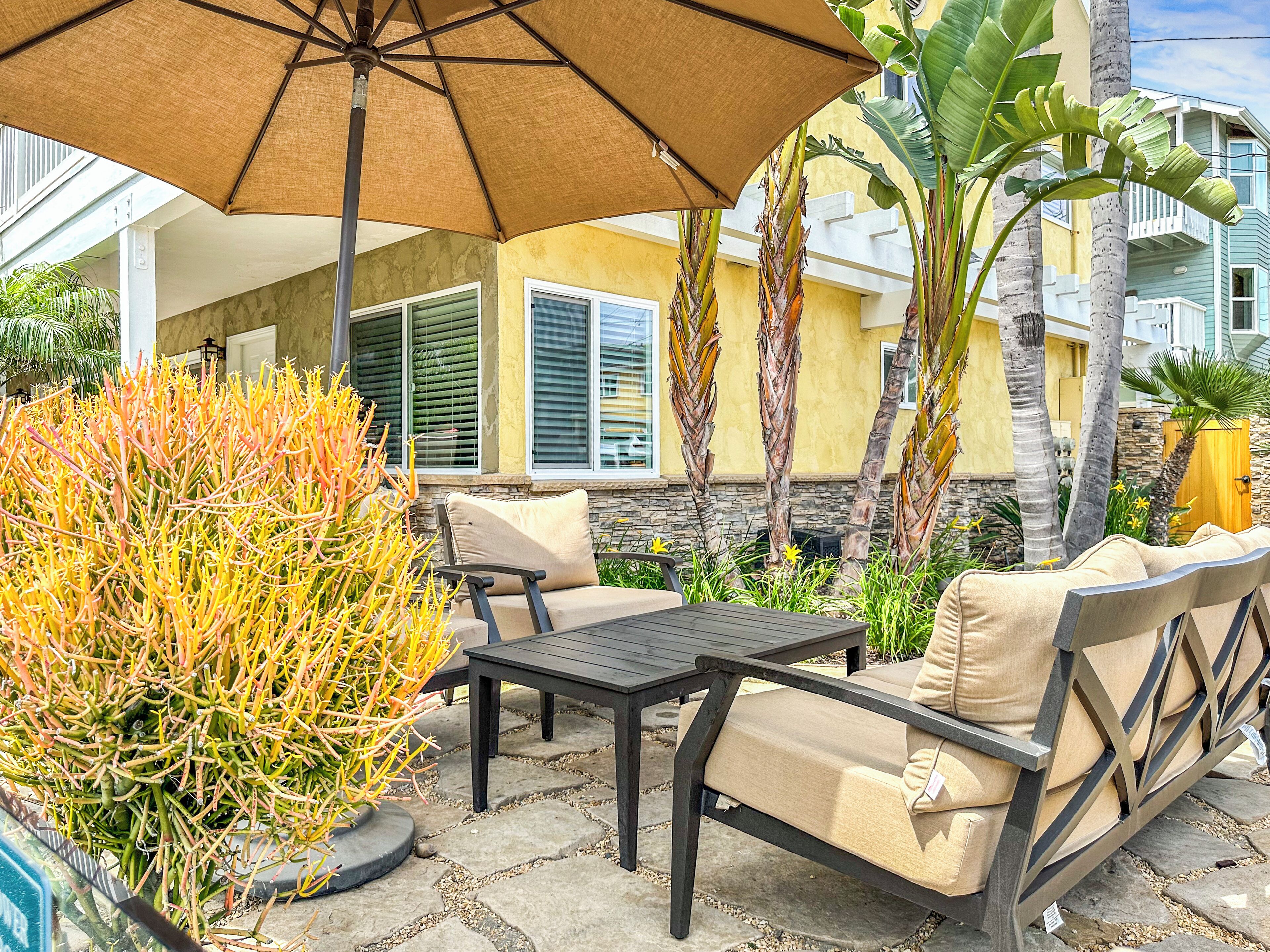 An outdoor patio area with a table and chairs, a large umbrella, and tropical plants.
