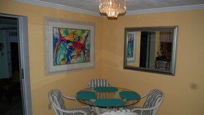 A dining area with a glass table surrounded by wicker chairs, a colorful abstract painting on the wall, and a mirror reflecting the room. - Old Florida Charm, quiet, close to beach and boat launch. (Englewood)