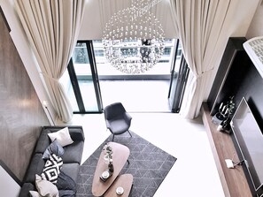 A modern living room with a sofa, a coffee table, and a chair. There is a large window with curtains, a chandelier, and a view of the outside. - Luxury Duplex at The Reach Titiwangsa (Kuala Lumpur)