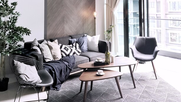 A modern living room with a herringbone-patterned wall, a grey sofa with patterned cushions, a coffee table, and a black armchair.