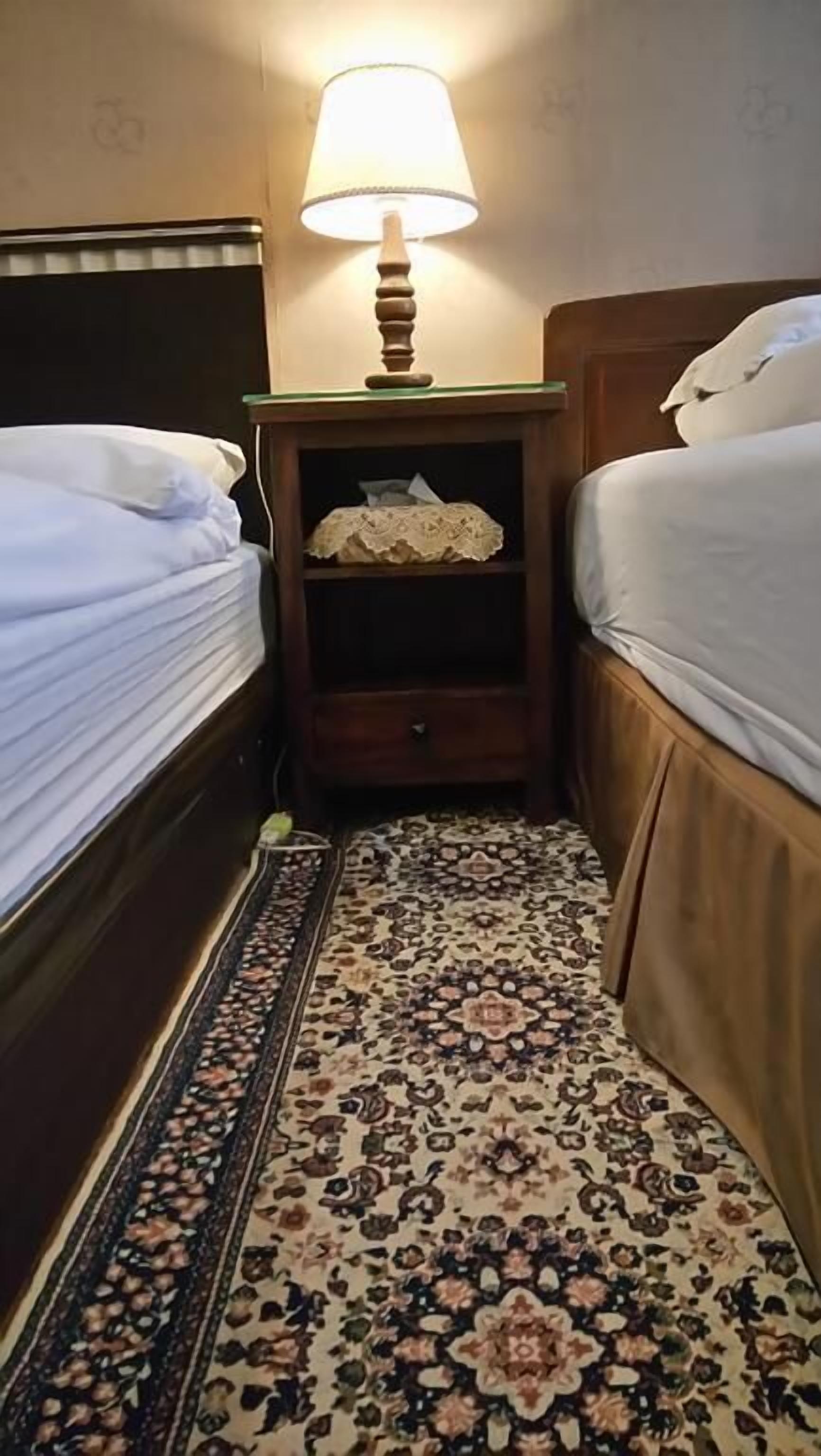 Ein Hotelzimmer mit zwei Einzelbetten, einem Nachttisch mit Lampe, einer kleinen Ablage mit Spitzenklöppel und einem gemusterten Teppich.