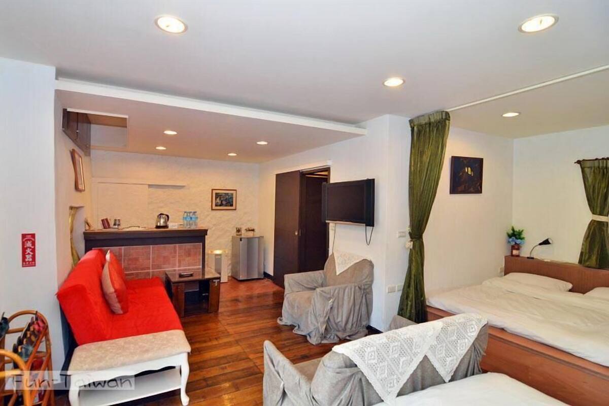 A hotel room with a bed, a seating area with a red chair, a small table, and a TV mounted on the wall.