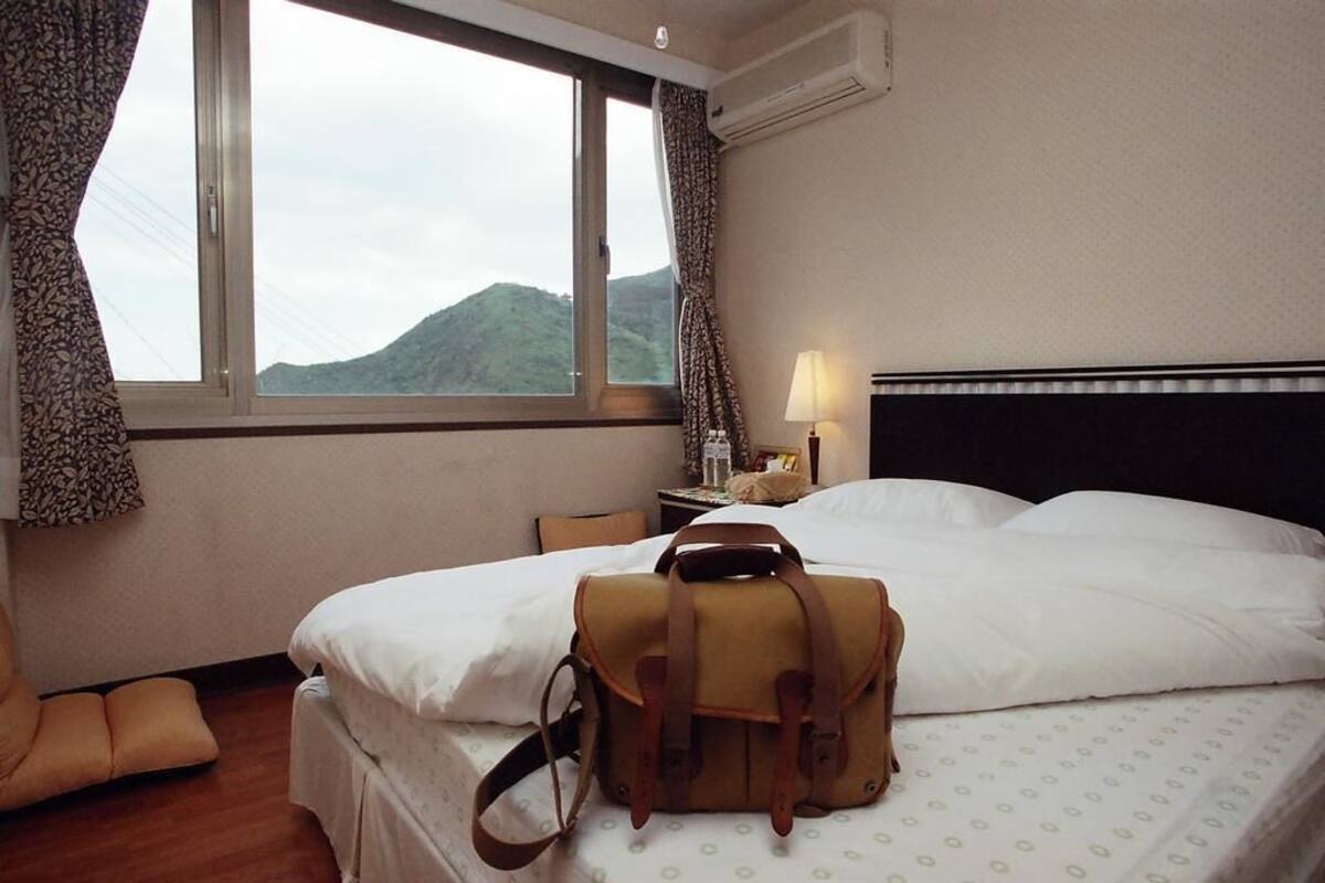 A hotel room with a bed, a nightstand, a lamp, and a view of mountains through the window.