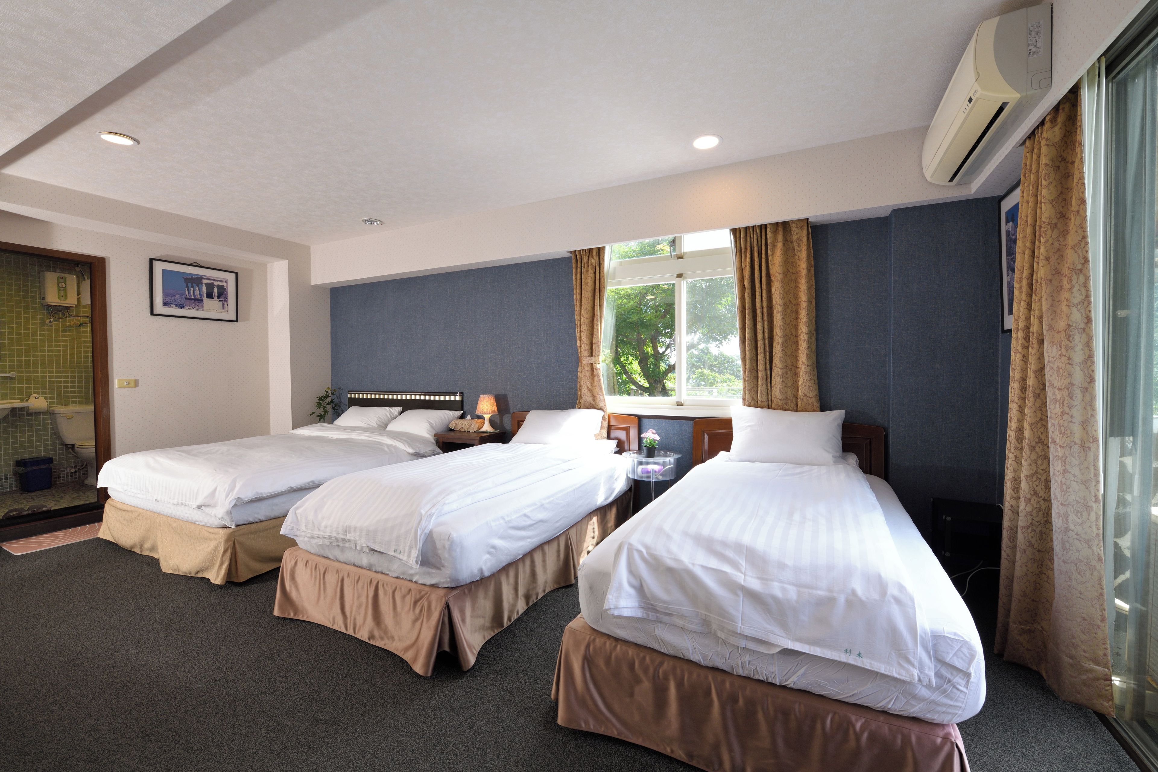 A hotel room with two single beds, a small table with a lamp, a window with curtains, and an air conditioning unit.
