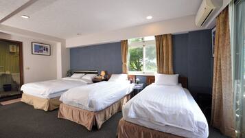 A hotel room with two single beds, a small table with a lamp, a window with curtains, and an air conditioning unit.