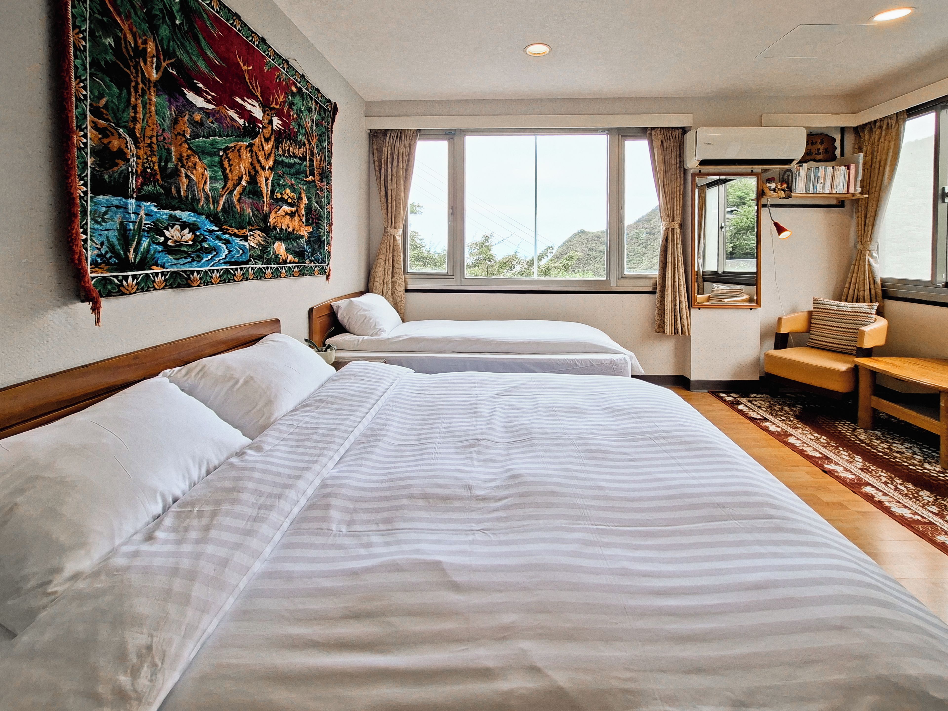 A hotel room with two beds, a large tapestry, a window with curtains, and a small table with a chair.