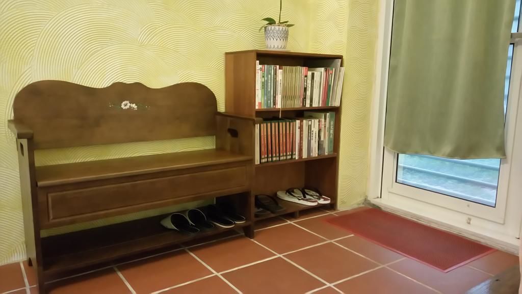 Um banco de madeira com prateleira embaixo, uma estante de livros e um par de sapatos no chão.