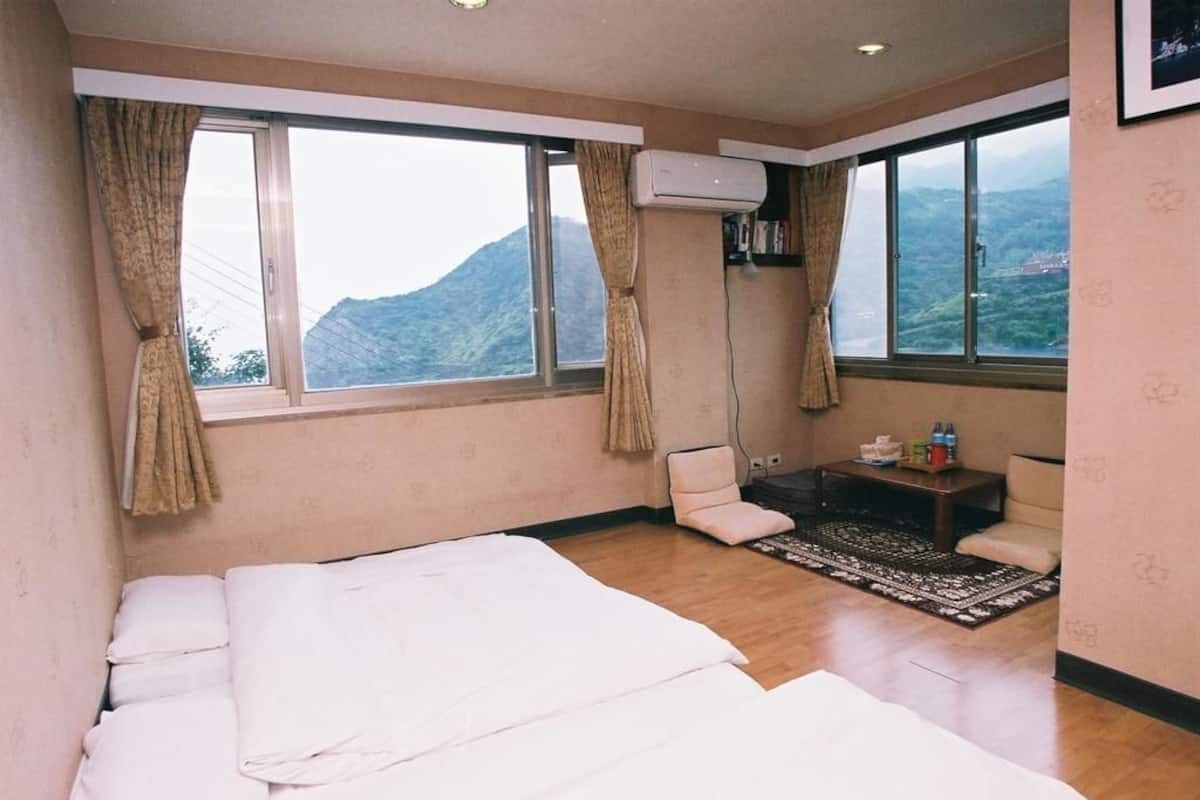 A hotel room with a large bed, two windows with curtains, a small table with items, a chair, and a view of mountains.