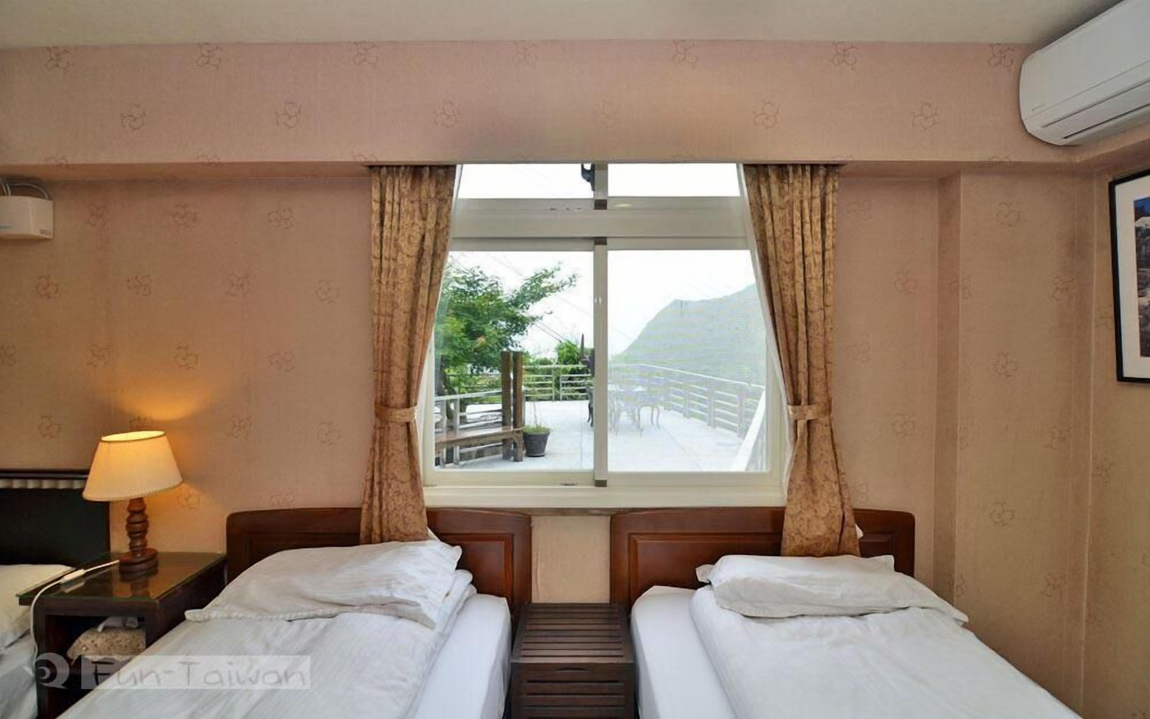 Une chambre d’hôtel avec deux lits, une fenêtre avec des rideaux, une table de chevet avec une lampe et un appareil de climatisation.