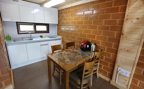 A compact kitchen with white cabinets, a sink, and a window above it. There's a dining table with chairs and a red kettle on it. - Jeju Starry Sky Pension (Seogwipo)