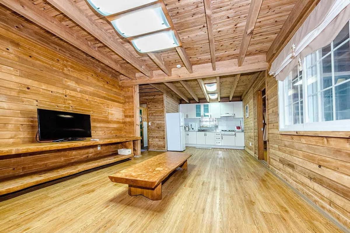 Interior de una cabaña de madera con televisor, banca de madera y zona de cocina.