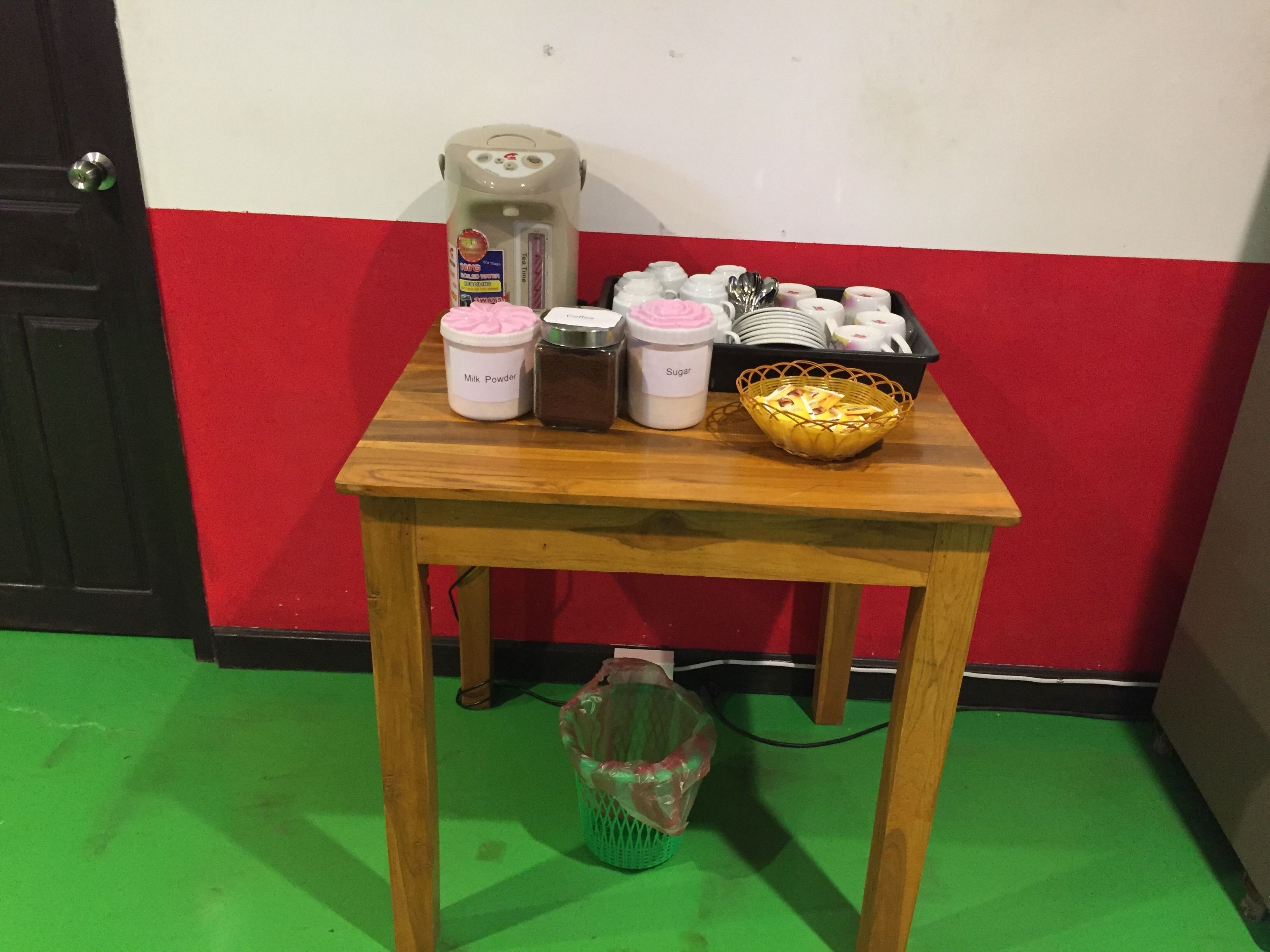 A small wooden table with kitchen items including a milk powder container, a sugar container, a coffee maker, and a basket of bread.