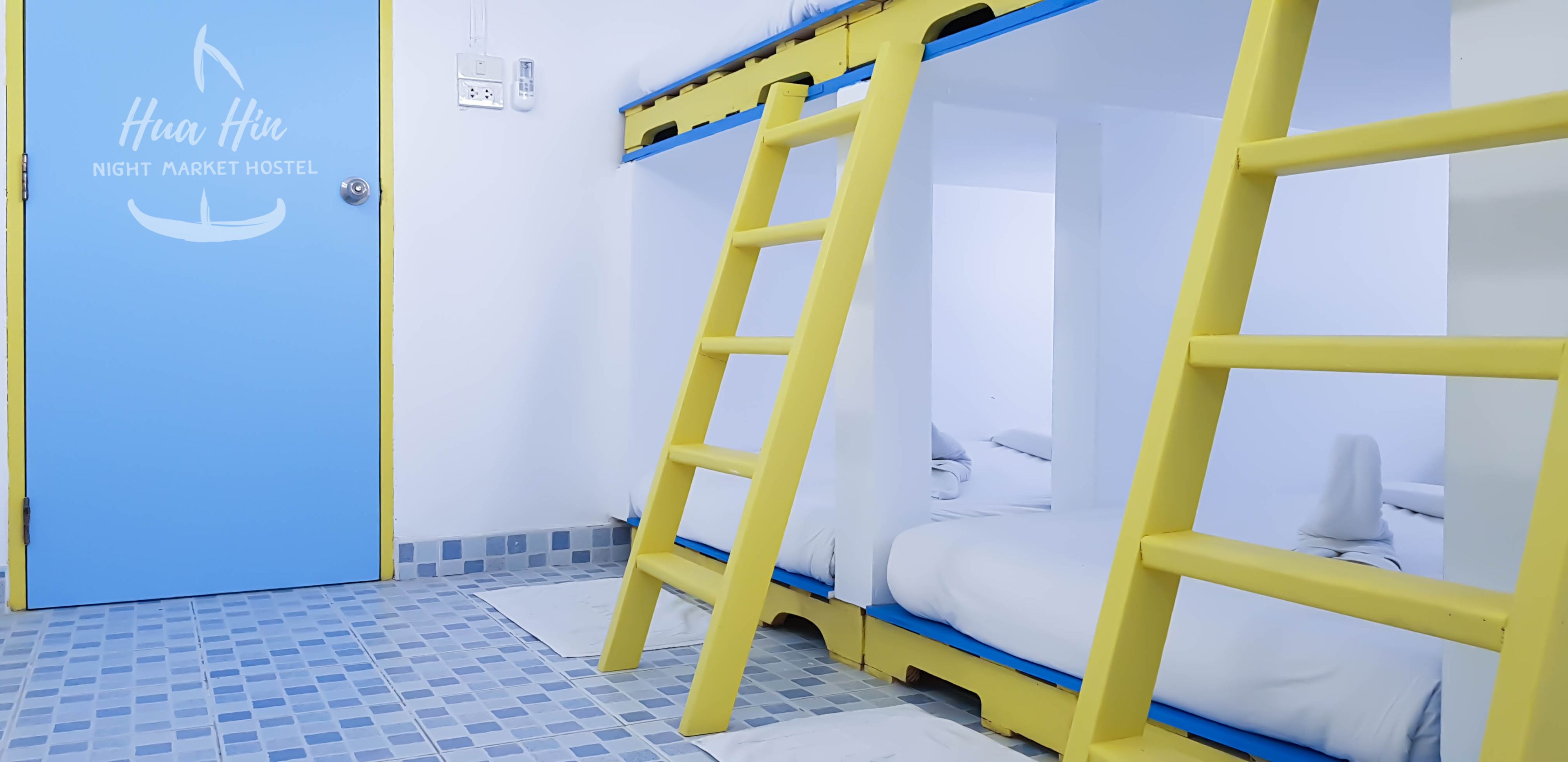 Habitación con literas, una escalera, una puerta azul con un logo y suelo de baldosas.