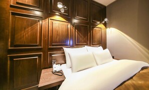 A hotel room with a wooden panel wall, a bed with white linens, a bedside table with a telephone, and a wall-mounted lamp. - A-VANT HOTEL (Cheonan)