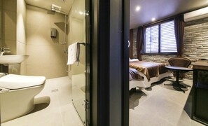 A modern bathroom with a glass shower enclosure, a white sink, and a toilet. The room features a bed with a brown headboard, a small round table, and a stone wall. - A-VANT HOTEL (Cheonan)