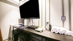 Une chambre d’hôtel équipée d’un bureau avec un téléviseur à écran plat, un ordinateur portable, une machine à café et une télécommande. On trouve également une serviette et un petit récipient blanc sur le bureau.