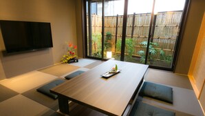 Una habitación con suelo de esteras de tatami, una mesa baja, cojines, un televisor de pantalla plana y vistas a una valla de bambú y un jardín.
